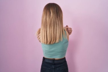 Blonde caucasian woman standing over pink background hugging oneself happy and positive from backwards. self love and self care 