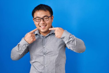 Mavi arka planda dikilen genç Çinli adam gülümsüyor ve parmaklarıyla ve ağzıyla işaret ediyor. diş sağlığı kavramı. 
