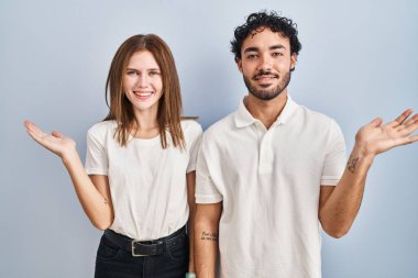 Gündelik giysiler giymiş genç bir çift neşeli bir şekilde ayakta duruyor ve avuç içi kameraya bakıyor.. 