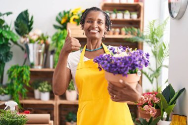 Çiçekçi dükkanında çalışan Afrikalı bir kadın elinde çiçeklerle gülümseyen mutlu ve olumlu bir şekilde başparmağıyla mükemmel ve onay işareti yapıyor. 