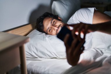 Genç İspanyol adam yatakta akıllı telefon kullanıyor.