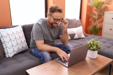 Orta yaşlı bir adam evdeki koltukta dizüstü bilgisayar kullanıyor.