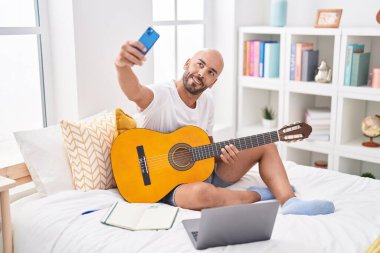 Young bald man playing classical guitar make selfie by smartphone at bedroom