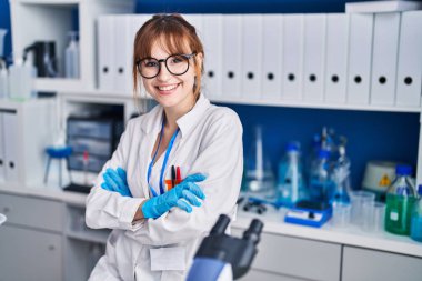 Genç bilim kadını gülümsüyor. Kollarını kavuşturup laboratuvarda duruyor.