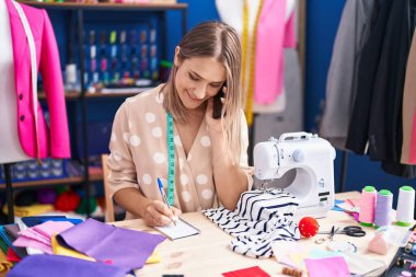Young caucasian woman tailor talking on smartphone writing on notebook at sewing studio