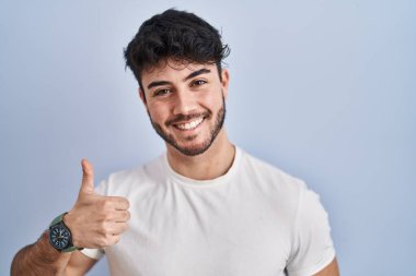 Hispanic man with beard standing over white background doing happy thumbs up gesture with hand. approving expression looking at the camera showing success. 