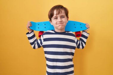 Küçük beyaz çocuk sarı arka planda kaykay tutuyor komik yüzlü yanakları şişiriyor. Ağzı havayla şişer, havayı yakalar.. 