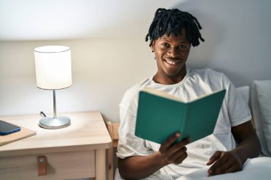 African american man reading book sitting on bed at bedroom