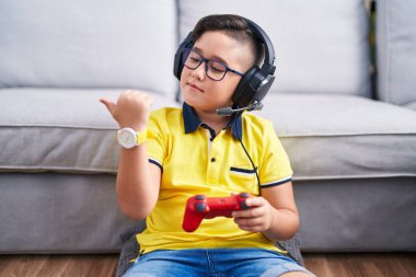 Video oyunu oynayan, kulaklık takan, gülümseyen ve baş parmağıyla yüzünü gösteren İspanyol bir çocuk.. 