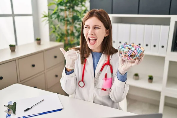 Joven Doctora Sosteniendo Caramelos Apuntando Con Pulgar Hacia Lado Sonriendo — Foto de Stock