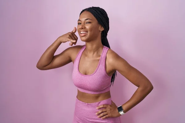 ピンクの背景にスポーツウェアを着たアフリカ系アメリカ人の女性は 電話で話すような手や指で携帯電話のジェスチャーをして笑っています コミュニケーションの概念 — ストック写真