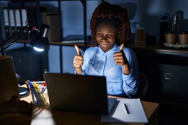 African woman working at the office at night success sign doing positive gesture with hand, thumbs up smiling and happy. cheerful expression and winner gesture.