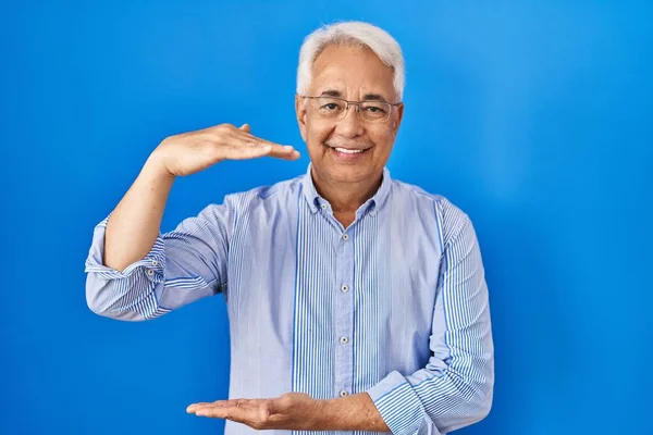 Hispanic senior man wearing glasses gesturing with hands showing big and large size sign, measure symbol. smiling looking at the camera. measuring concept.