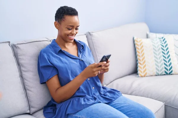 アフリカ系アメリカ人の女性が自宅のソファに座ってスマートフォンを使って — ストック写真