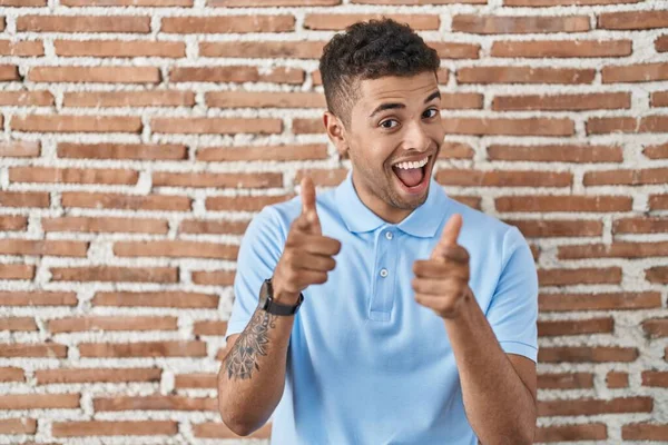ブラジルの若い男がレンガの壁の上に立ってカメラに指を指して幸せと面白い顔 良いエネルギーと振動は — ストック写真