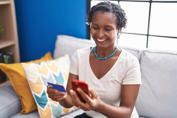 中年アフリカ系アメリカ人女性を使用してスマートフォンやクレジットカードを使用してソファに家に座って — ストック写真