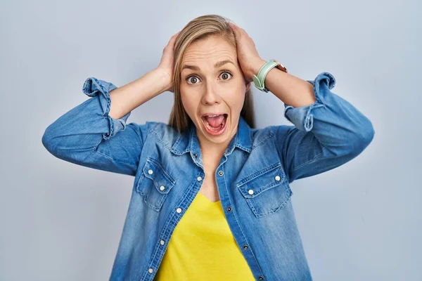 Young blonde woman standing over blue background crazy and scared with hands on head, afraid and surprised of shock with open mouth