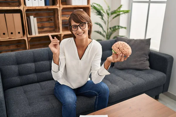 Brunette woman working at therapy office holding brain smiling happy pointing with hand and finger to the side