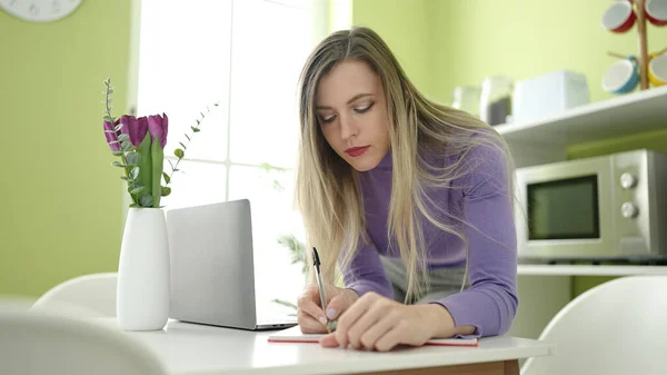 年轻的金发女人在家里用笔记本电脑在笔记本上写字 — 图库照片