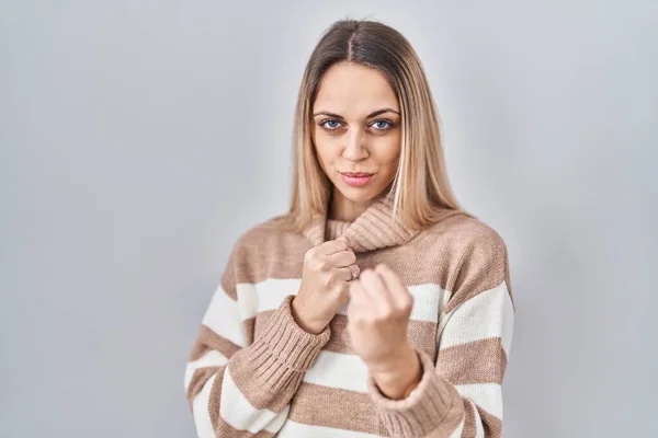 Young blonde woman wearing turtleneck sweater over isolated background ready to fight with fist defense gesture, angry and upset face, afraid of problem