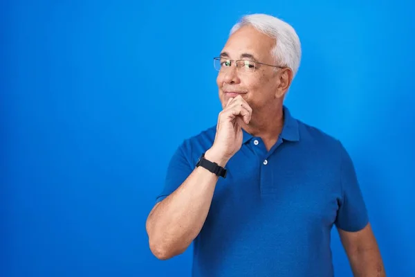 Middle age man with grey hair standing over blue background looking confident at the camera with smile with crossed arms and hand raised on chin. thinking positive.
