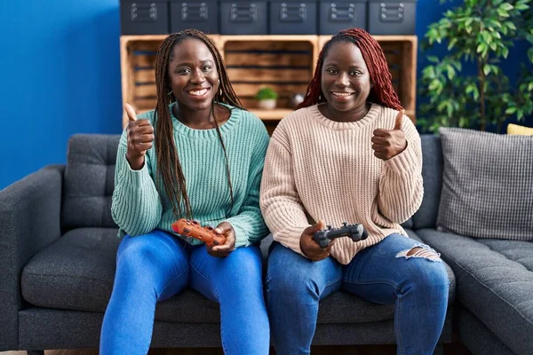 两名玩电子游戏的非洲女人拿着控制器开心而积极的笑着 竖起大拇指做得很好 并签了字 — 图库照片
