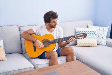 Young hispanic man playing classical guitar sitting on sofa at home