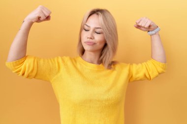 Sarı kazak giyen genç beyaz kadın, gururla gülümseyen kol kaslarını gösteriyor. fitness konsepti. 