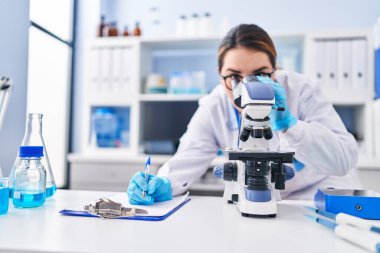 Young beautiful hispanic woman scientist using microscope writing on document at laboratory