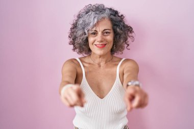 Pembe arka planda gri saçlı orta yaşlı bir kadın seni ve kamerayı işaret ediyor. 