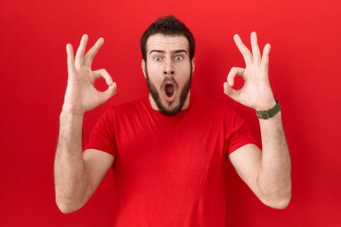 Günlük kırmızı tişört giyen genç İspanyol adam şaşırmış ve şok olmuş bir şekilde parmaklarıyla onay sembolü yapıyor. çılgın ifade 