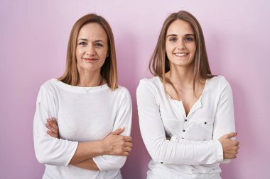 Orta yaşlı anne ve küçük kızı pembe arka planda durup kollarını kavuşturup kameraya bakarken gülümsüyor. pozitif kişi. 