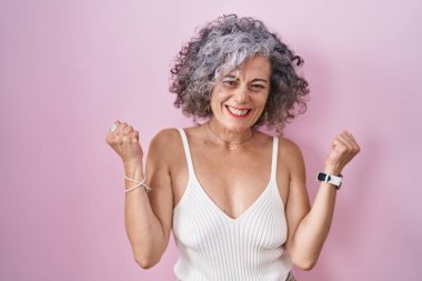 Pembe arka planda duran gri saçlı orta yaşlı kadın kolları havada ve gözleri kapalı bir şekilde başarıyı kutluyor ve şaşırıyor. Kazanan konsept. 