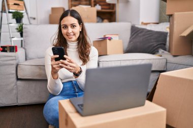 Young beautiful hispanic woman using laptop and smartphone sitting on floor at new home