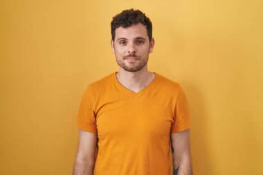 Young hispanic man standing over yellow background relaxed with serious expression on face. simple and natural looking at the camera. 