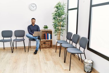 Genç İspanyol adam bekleme odasında otururken dizüstü bilgisayar kullanıyor.