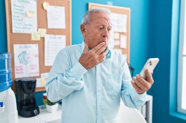 Ofiste çalışan gri saçlı son sınıf öğrencisi. Akıllı telefonuyla telefon görüşmesi yapıyor. Ağzı elle kapatıyor, şok geçiriyor ve hata yapmaktan korkuyor. şaşırmış ifade 