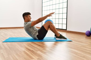 Genç İspanyol adam gülümsüyor. Spor merkezinde top kullanarak karın kasları çalıştırıyor.