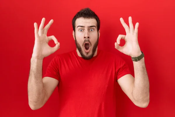 カジュアルな赤いTシャツを着た若いヒスパニック系の男性は驚き 指でOk承認シンボルをやってショックを受けました クレイジーな表現 — ストック写真