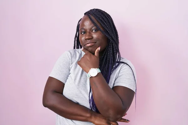 年轻的非洲女人站在粉红的背景上 自信地看着摄像机 双臂交叉 双手托着下巴微笑 积极地思考 — 图库照片