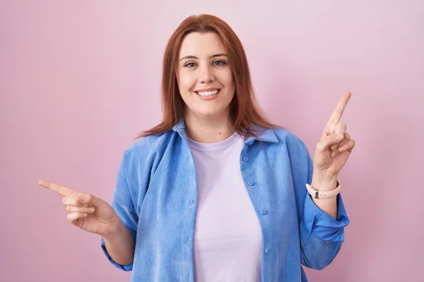 ピンクの背景に立っている赤い髪をした若いヒスパニック系の女性は 異なる方向への指で自信を持って指して笑顔 広告のコピースペース — ストック写真