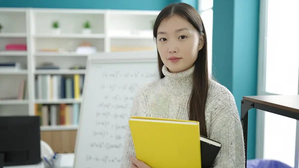 Young chinese woman student holding books standing with relaxed expression at library university