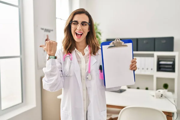 Young Doctor Woman Holding Clipboard Pointing Thumb Side Smiling Happy — Stock Photo, Image