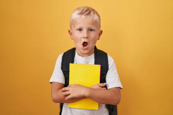 学生のバックパックを着て 本を持っている小さな白人の男の子は怖がって 驚きのために口を開けて驚いて 不信の顔 — ストック写真