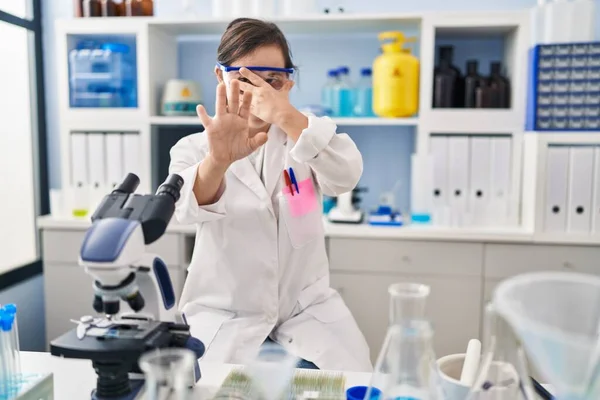 Hispanic girl with down syndrome working at scientist laboratory covering eyes with hands and doing stop gesture with sad and fear expression. embarrassed and negative concept.