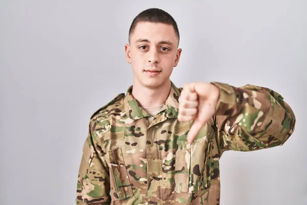 Young man wearing camouflage army uniform looking unhappy and angry showing rejection and negative with thumbs down gesture. bad expression.