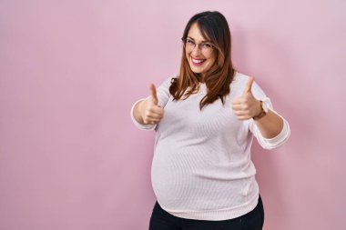 Pembe arka planda duran hamile bir kadın elleriyle olumlu bir jest yapmayı onaylıyor. Gülümsüyor ve başarı için mutlu. kazanan jest. 