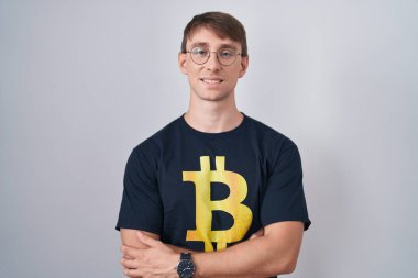 Bitcoin tişört giyen beyaz sarışın adam kollarını kavuşturup kameraya bakarken gülümsüyor. pozitif kişi. 