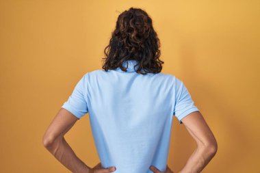 Young hispanic man standing over yellow background standing backwards looking away with arms on body 