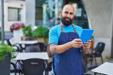 Young bald man waiter smiling confident using touchpad at coffee shop terrace